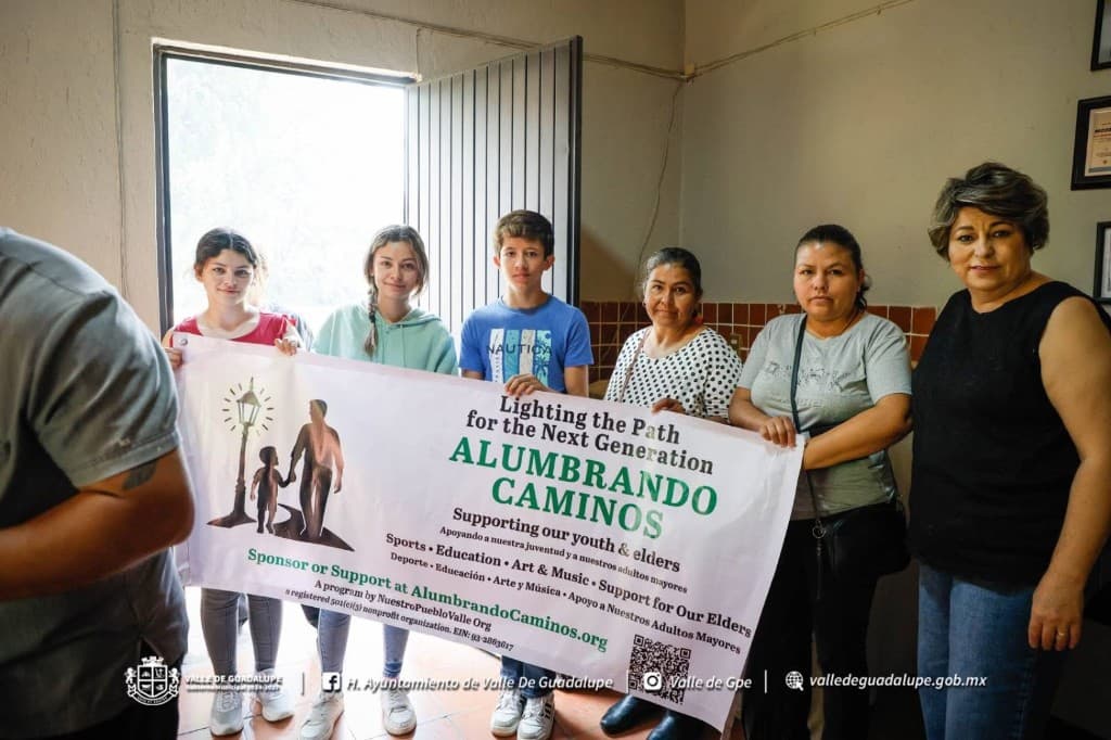 Volunteers holding the Alumbrando Caminos banner indoors — supporting youth, elders, sports, education, and the arts.